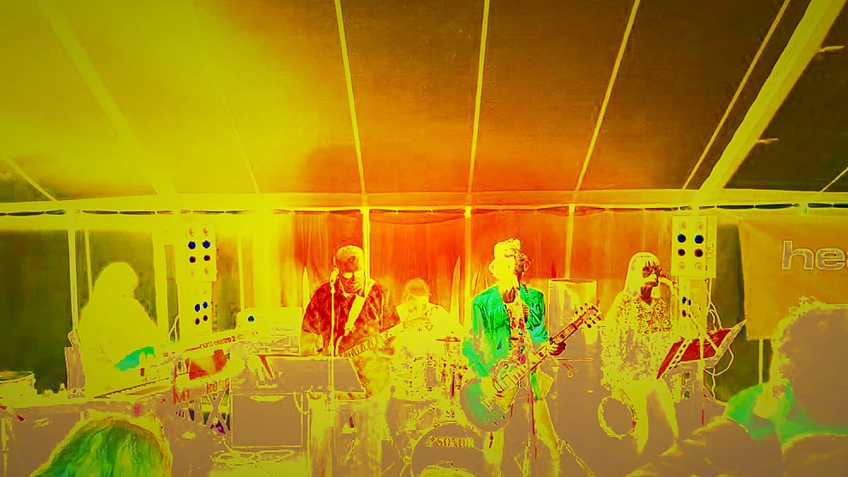 St Johns Wood Affair performing live under blue stage lighting inside a tent, keyboards and guitars upfront, crowd silhouettes in foreground.